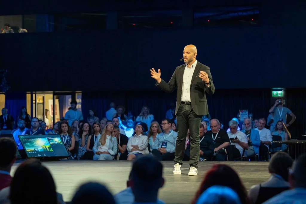 A man in a black suit and white shirt giving a presentation on stage in front of an audience at a conference or event.