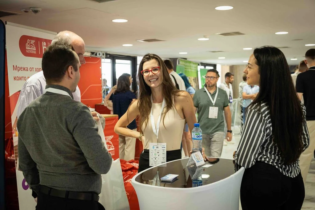 A group of four people, three women and one man, engaged in conversation at a professional networking event or conference. They are standing around a table with promotional materials, and there are other attendees in the background.