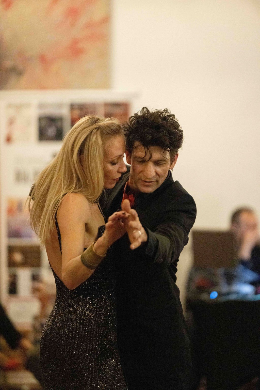 A man and woman dancing closely together at a social event, with the woman in a sparkly black dress and the man in a black outfit, both focused on their dance.