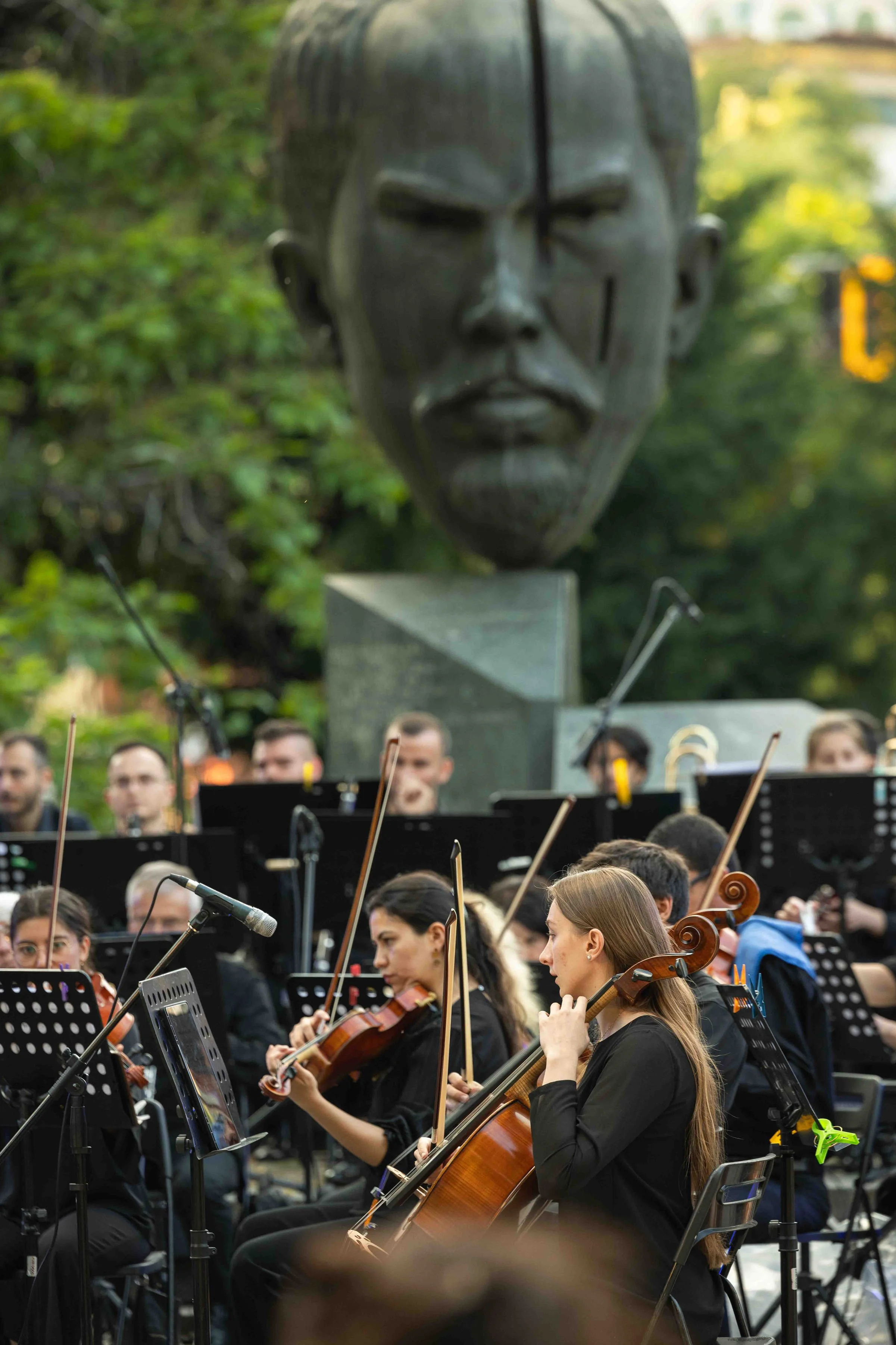 An outdoor orchestra performance with musicians playing string instruments such as violins and cellos, seated in front of a large sculpture of a person's face with mystical markings, set amidst lush greenery.
