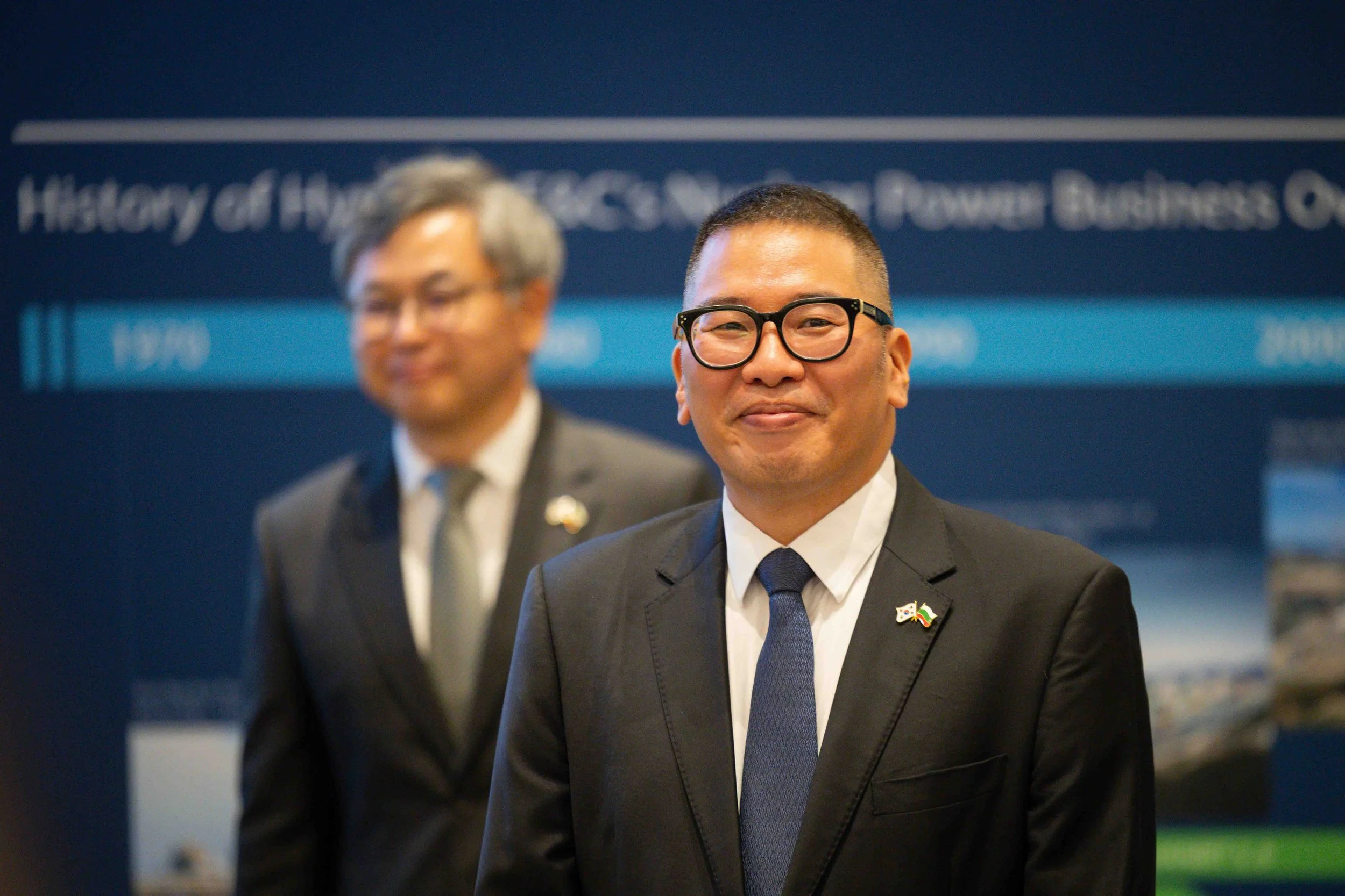 A man wearing glasses in a black suit and tie, smiling at the camera, with a South African flag pin on his lapel, standing in front of a blue background with blurred text and images. Another man in a suit and tie stands slightly out of focus behind h