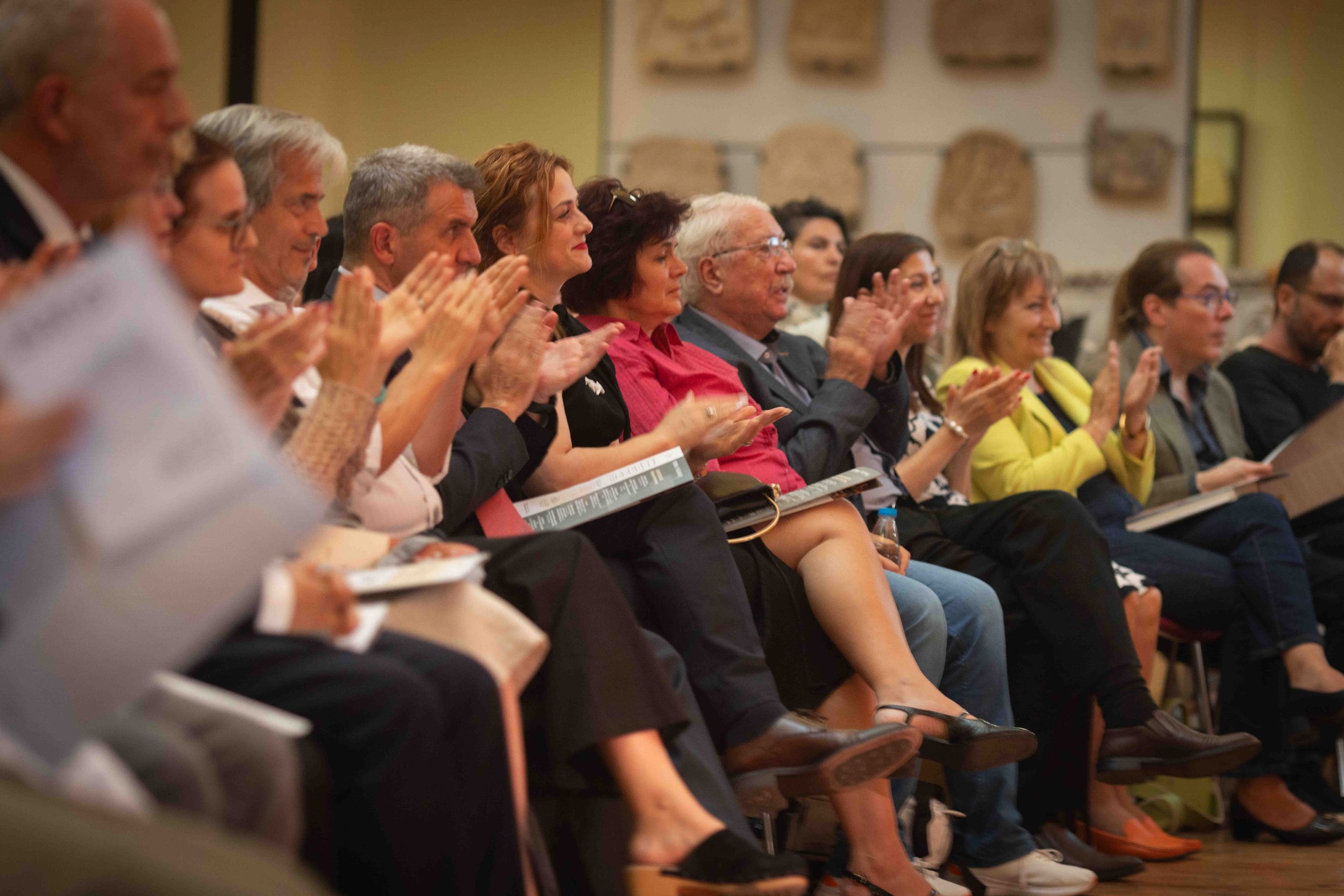 Audience members seated in a row, clapping and watching an event, with some holding printed programs, in an indoor setting.