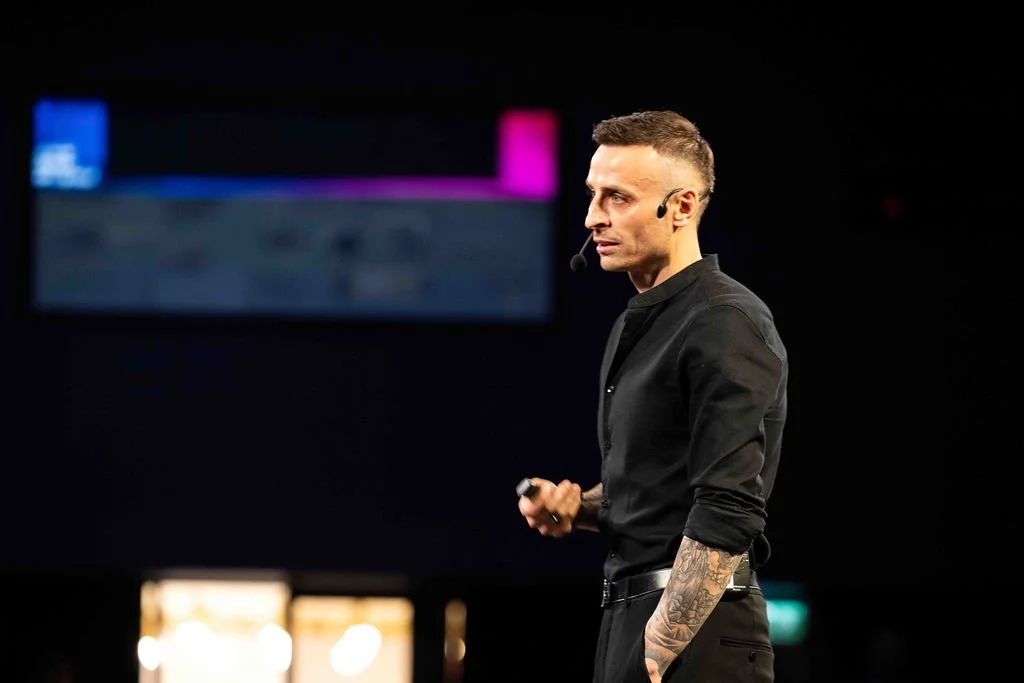 A man standing on stage, speaking into a microphone headset, wearing all black with tattoos visible on his left arm, with a large screen in the background displaying a blurred presentation.