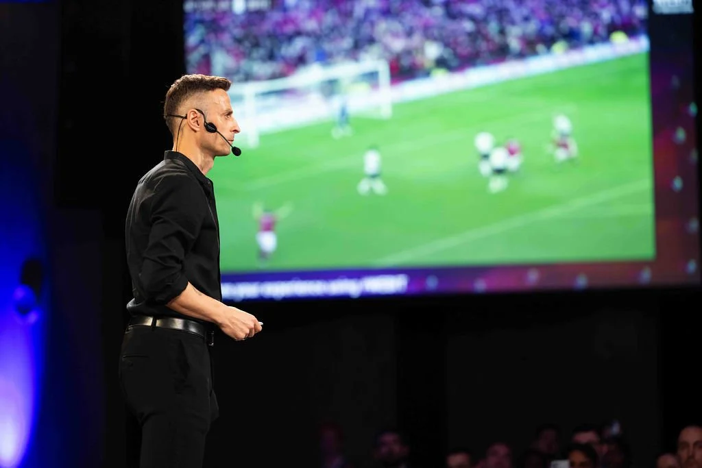 A man in black clothing with a headset microphone faces a large screen displaying a blurry soccer game.