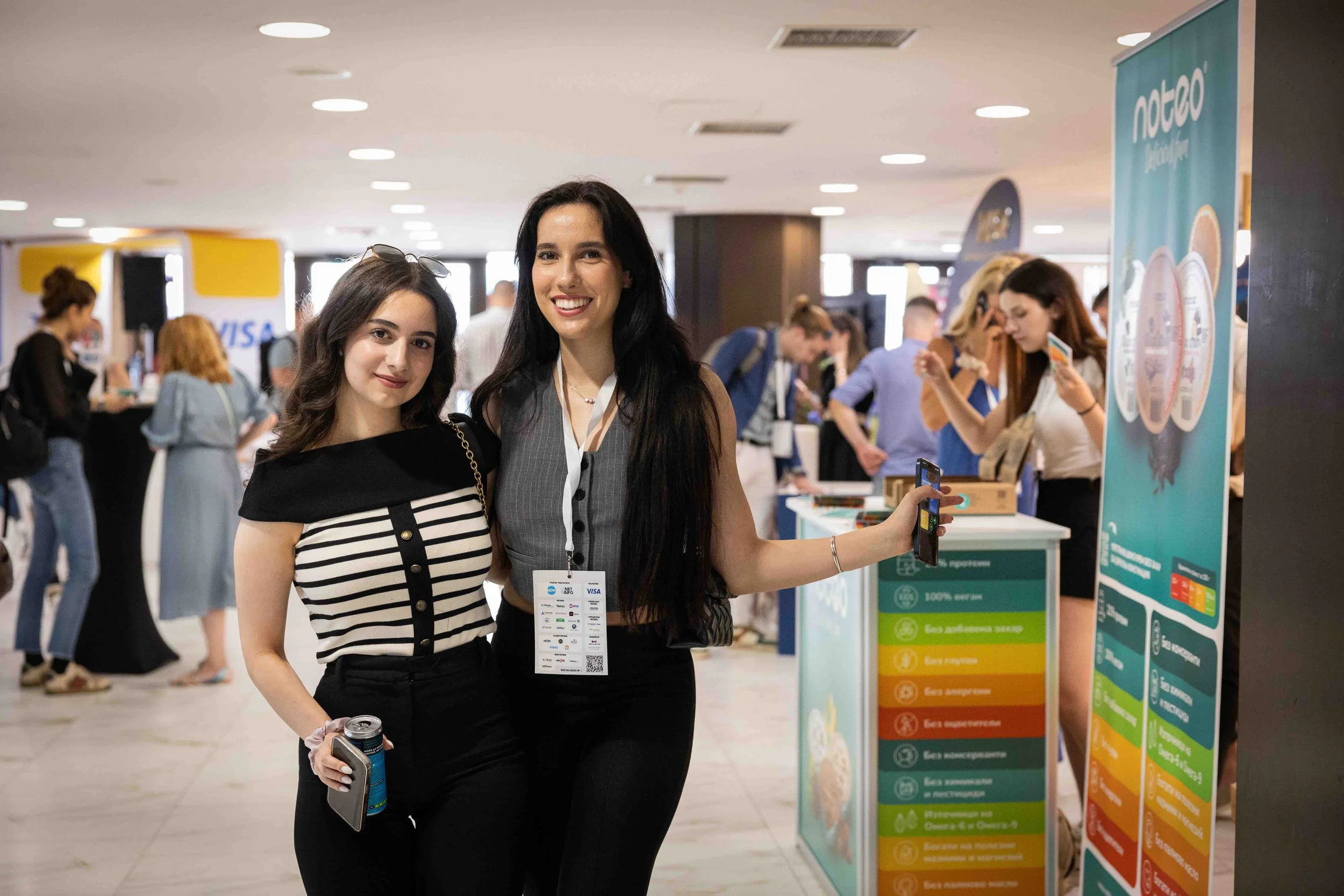 Two smiling attendees posing at the Webit 2025 expo zone, surrounded by booths and networking participants.