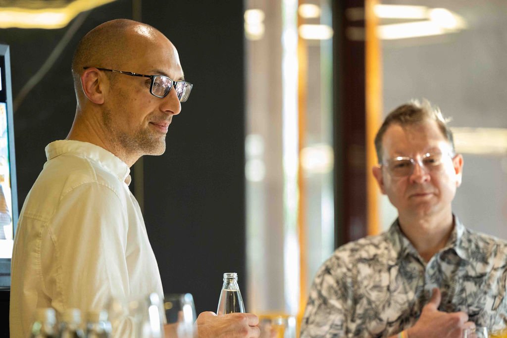 Two men with glasses in a casual setting, one holding a bottle, in a well-lit room with large windows.