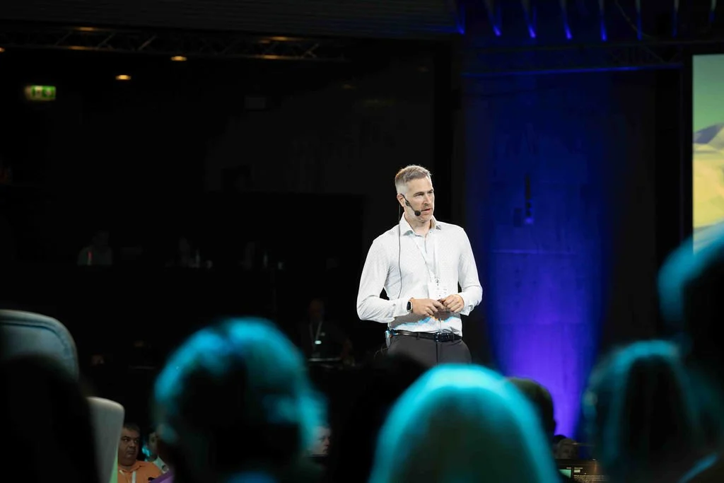 A man giving a presentation on a stage with dark background and purple lighting, wearing a white shirt and headset, in front of an audience.