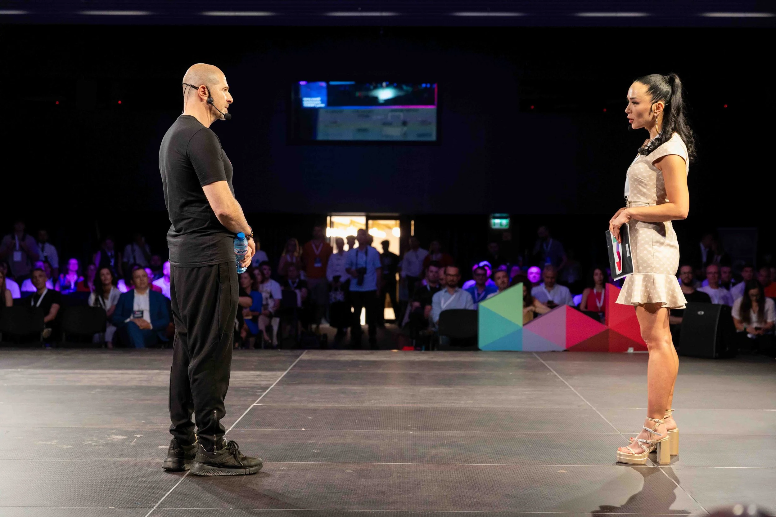 A man and woman are standing on a stage facing each other in front of a seated audience at a conference or event.