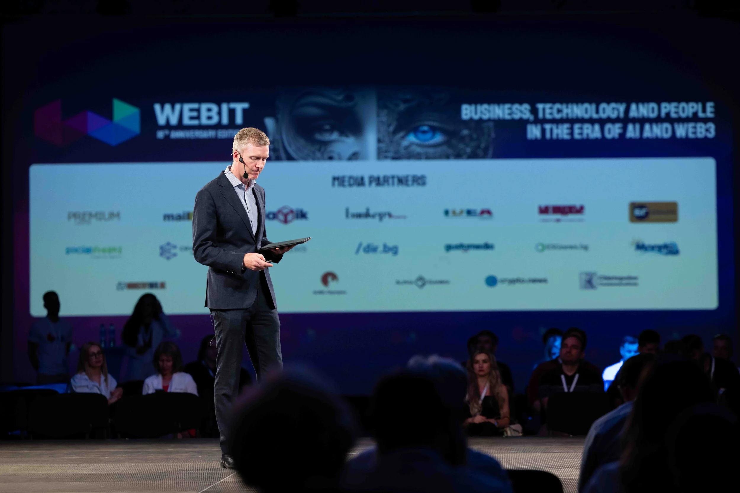 A man in a suit with a headset holding a tablet, standing on a stage at a conference. Behind him, a large screen displays the WEBIT 9th anniversary event with media partners listed, and the theme about business, technology, and people in the era of AI and Web3.