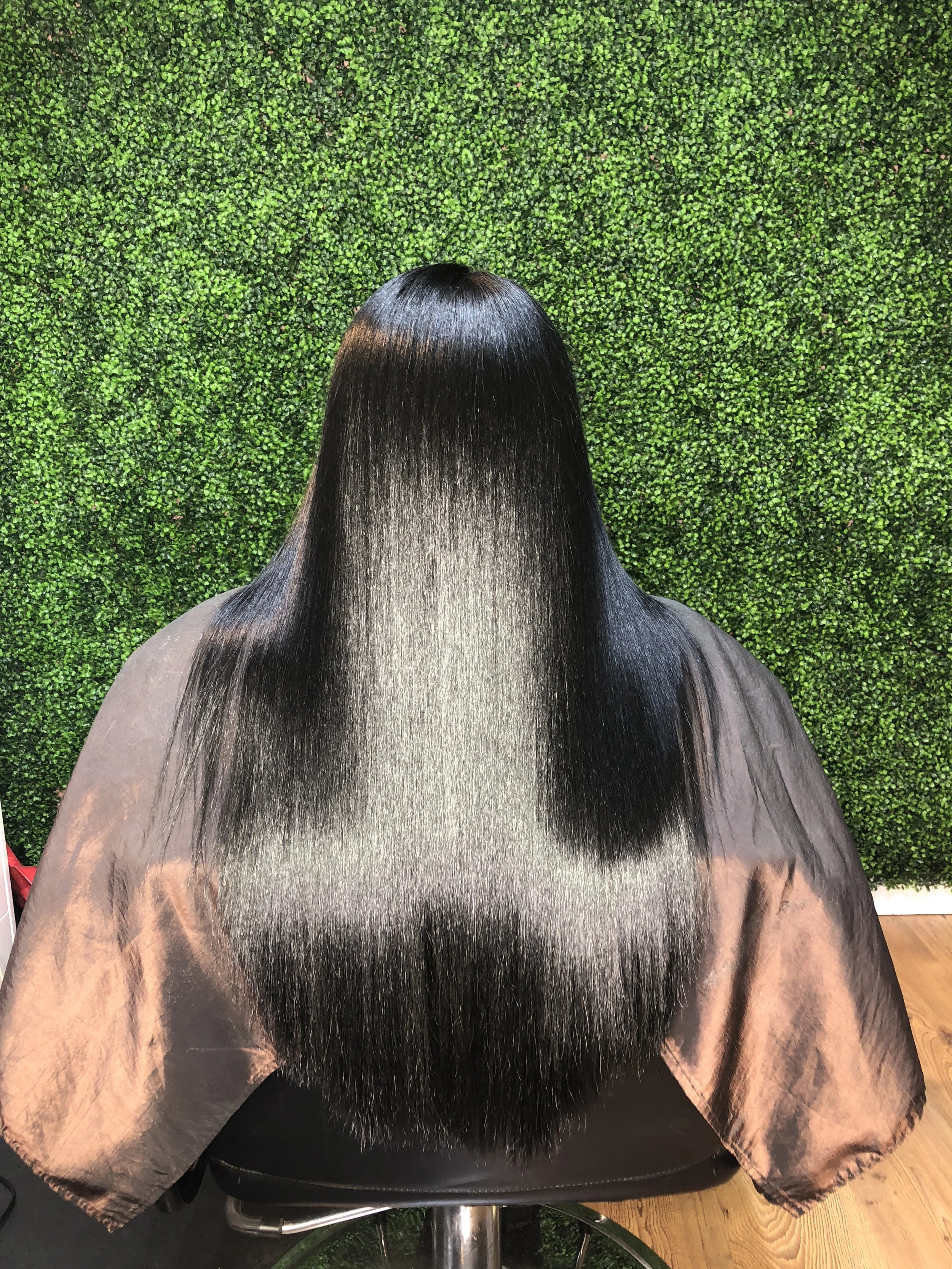 Back of a woman with long, straight, shiny black hair sitting in a salon chair in front of a green leafy wall.