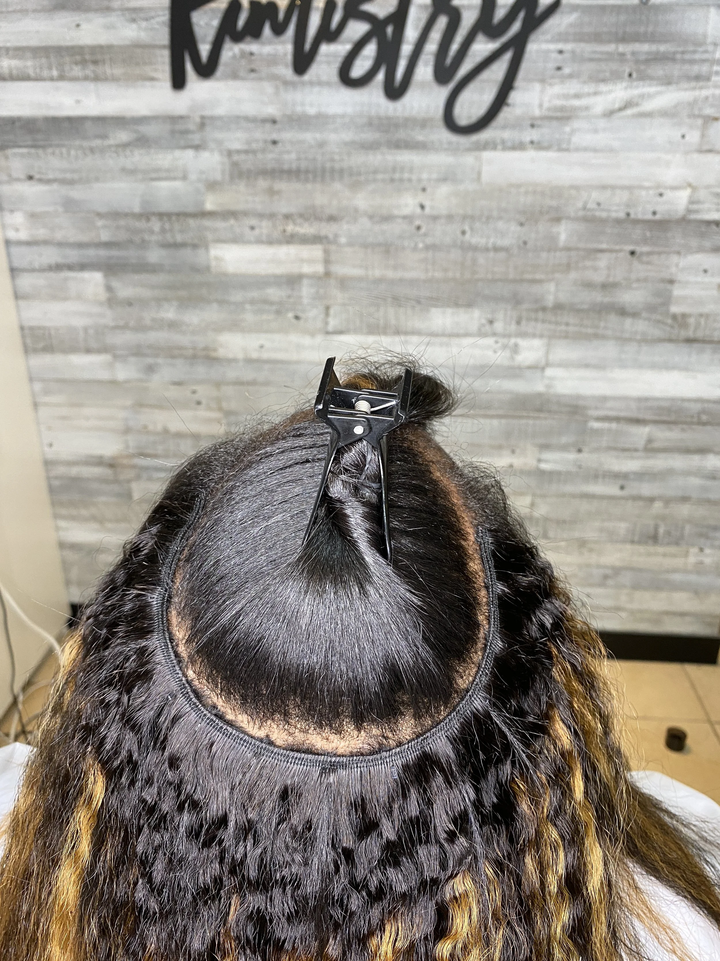 Top view of a person with curly hair and blonde highlights getting a hair weave or extensions applied, with a hair clip holding the hair in place, in a salon setting.