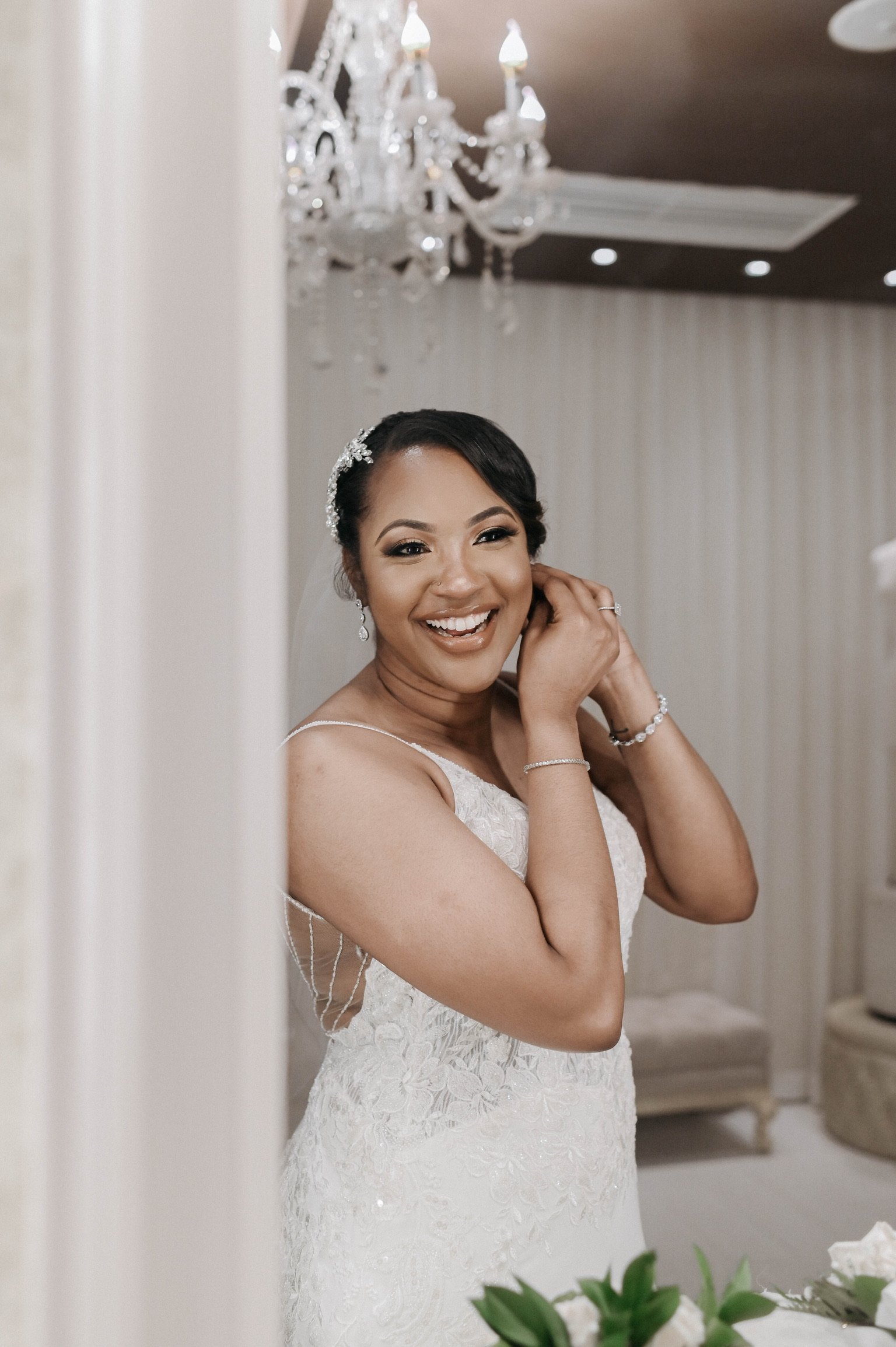 A bride in a wedding dress is smiling while getting ready, adjusting her earring in a room decorated with a chandelier.
