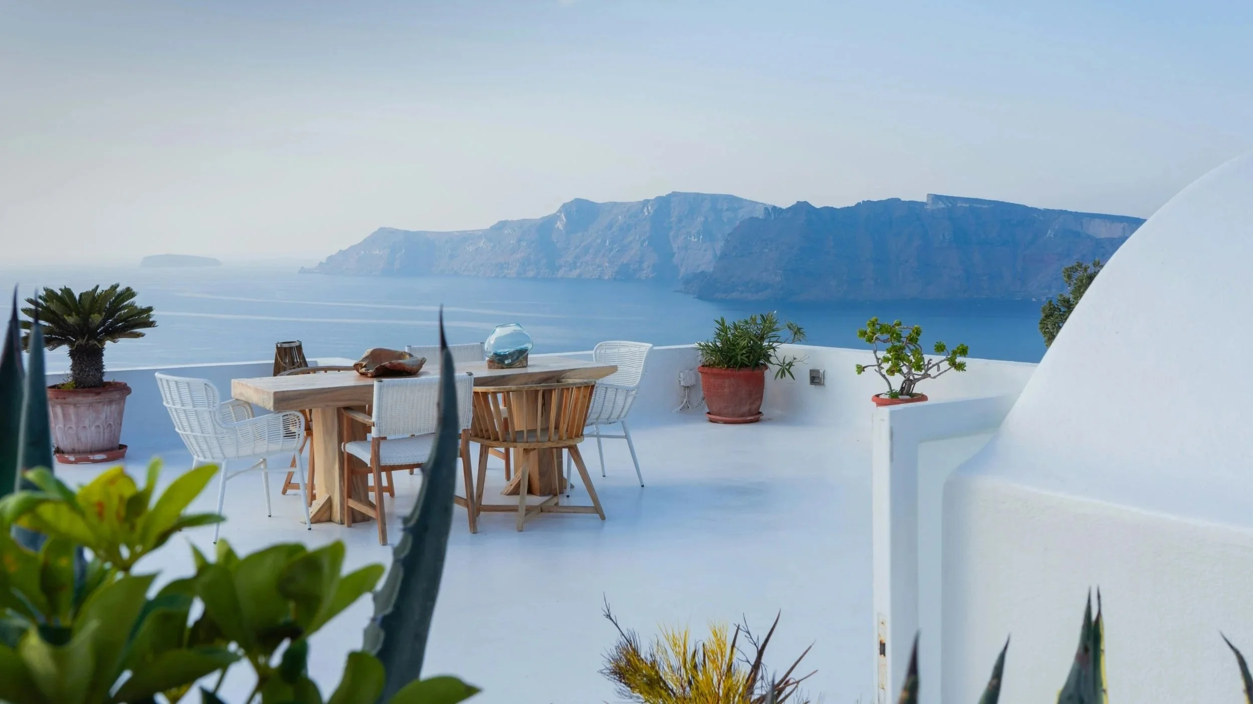 A scenic outdoor terrace with white walls and furniture, decorated with potted plants, overlooking a large body of water and mountainous coastline in the distance.