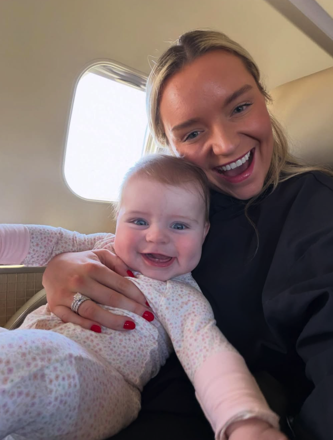 A woman and a baby smiling and taking a selfie inside an airplane. The woman has blonde hair and is wearing a black top. The baby has light hair and is wearing a light-colored outfit with small patterns.