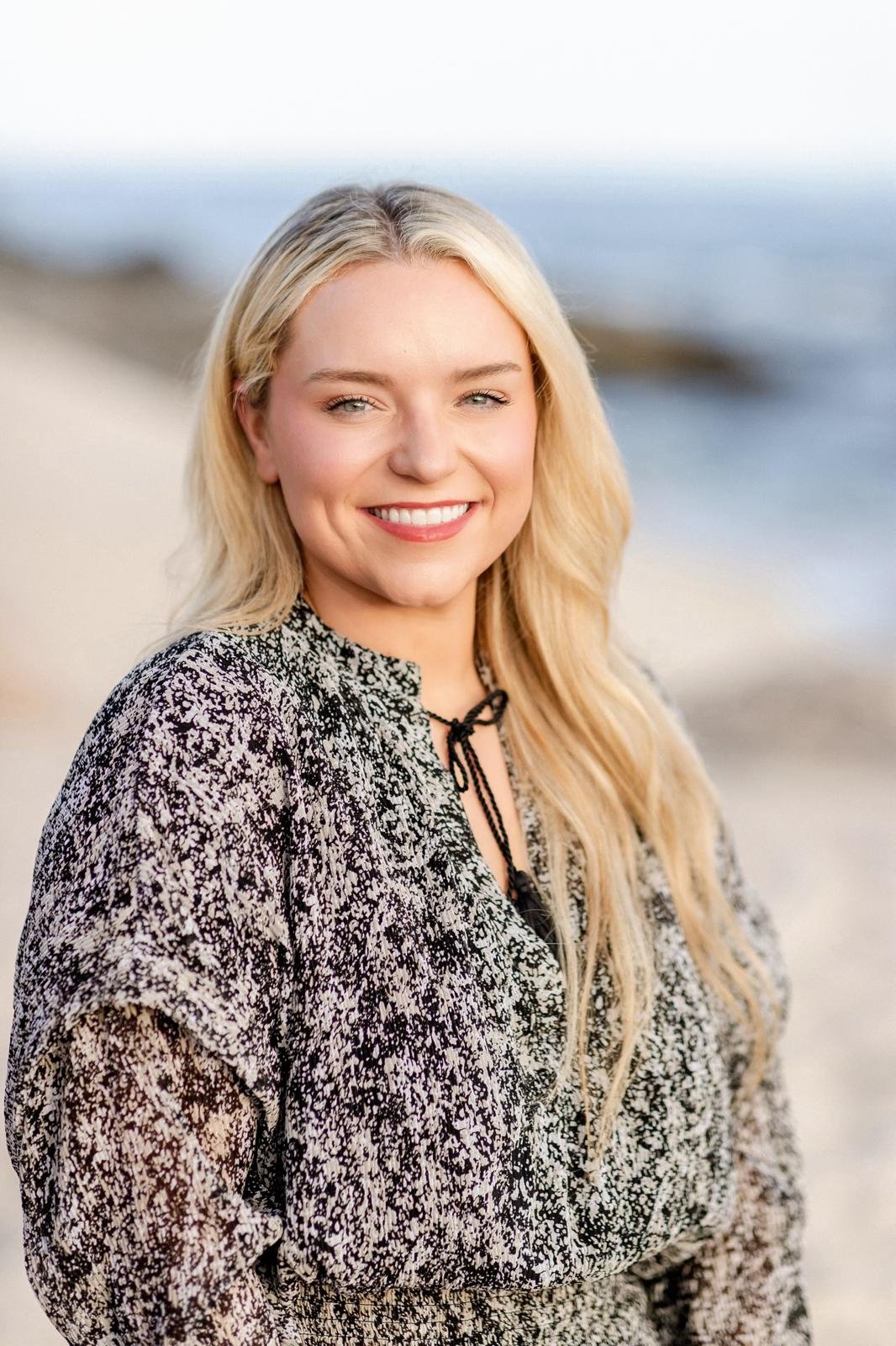 A young woman with long blonde hair and blue eyes smiling at the camera, standing on a beach with sand and water in the background.