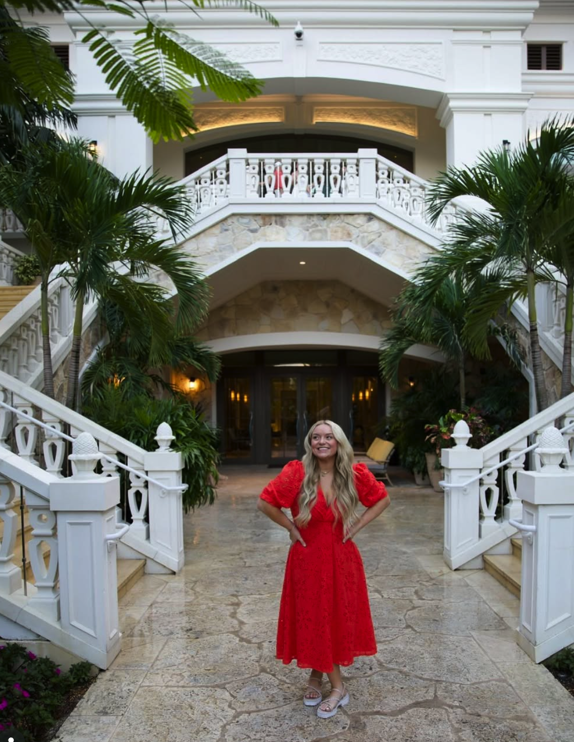 A woman in a red dress standing in front of a large white luxury house with palm trees and lush greenery.