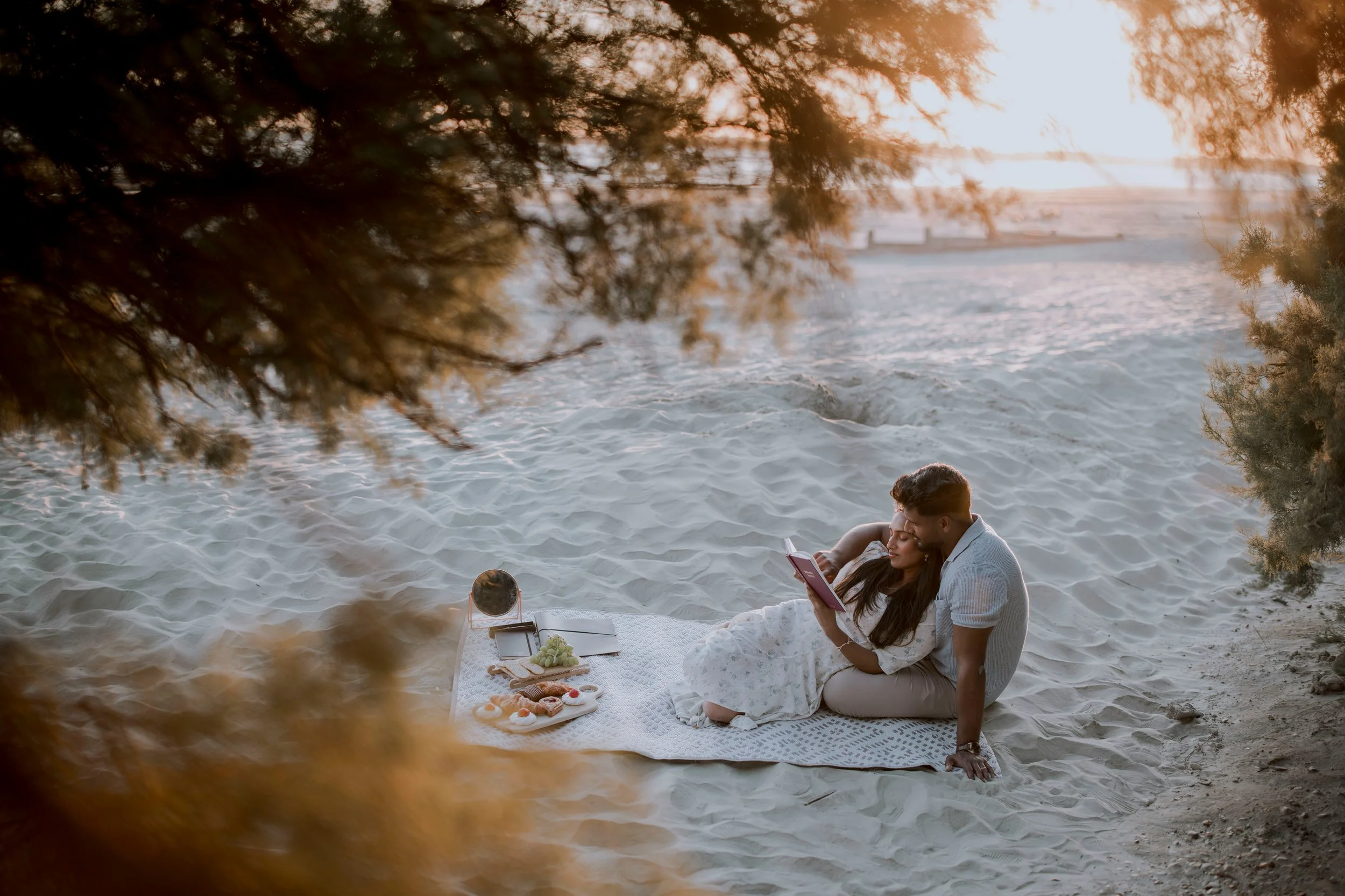 A couple sitting on a blanket on the sand at the beach, looking at a book, with a picnic setup nearby, during sunset and framed by tree branches.