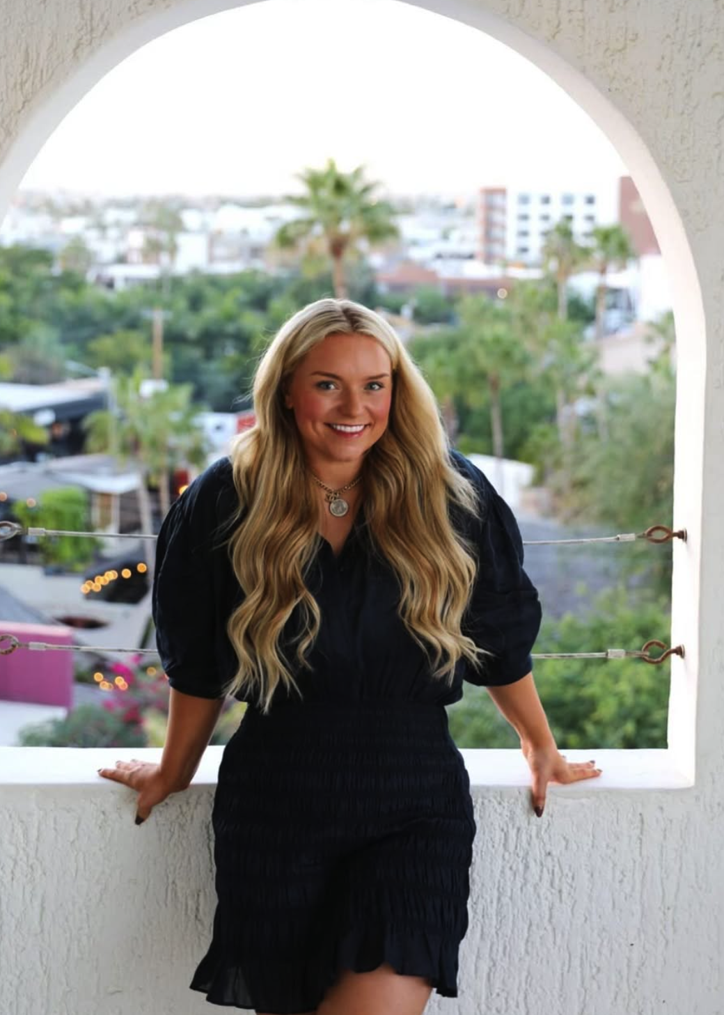 A woman with long blonde wavy hair smiling and leaning on a balcony railing, wearing a black dress, with a cityscape and palm trees in the background.