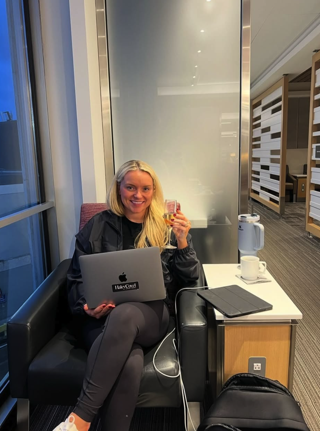 A woman with long blonde hair, smiling, sitting on a black leather armchair in an airport lounge. She holds a glass of white wine in one hand and a closed laptop on her lap. There is a small table beside her with a white mug, a water pitcher, and a black tablet. The background shows large windows and airport furniture.