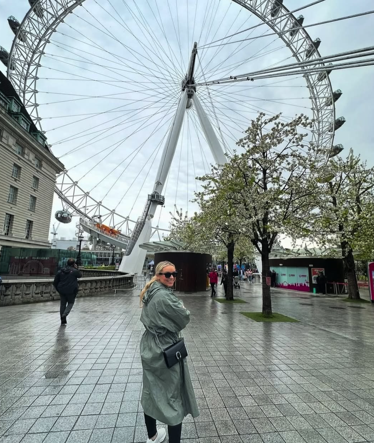 A woman smiling and wearing sunglasses stands on a wet pavement near blooming trees and a large Ferris wheel in an outdoor city setting. Several people are walking in the background.