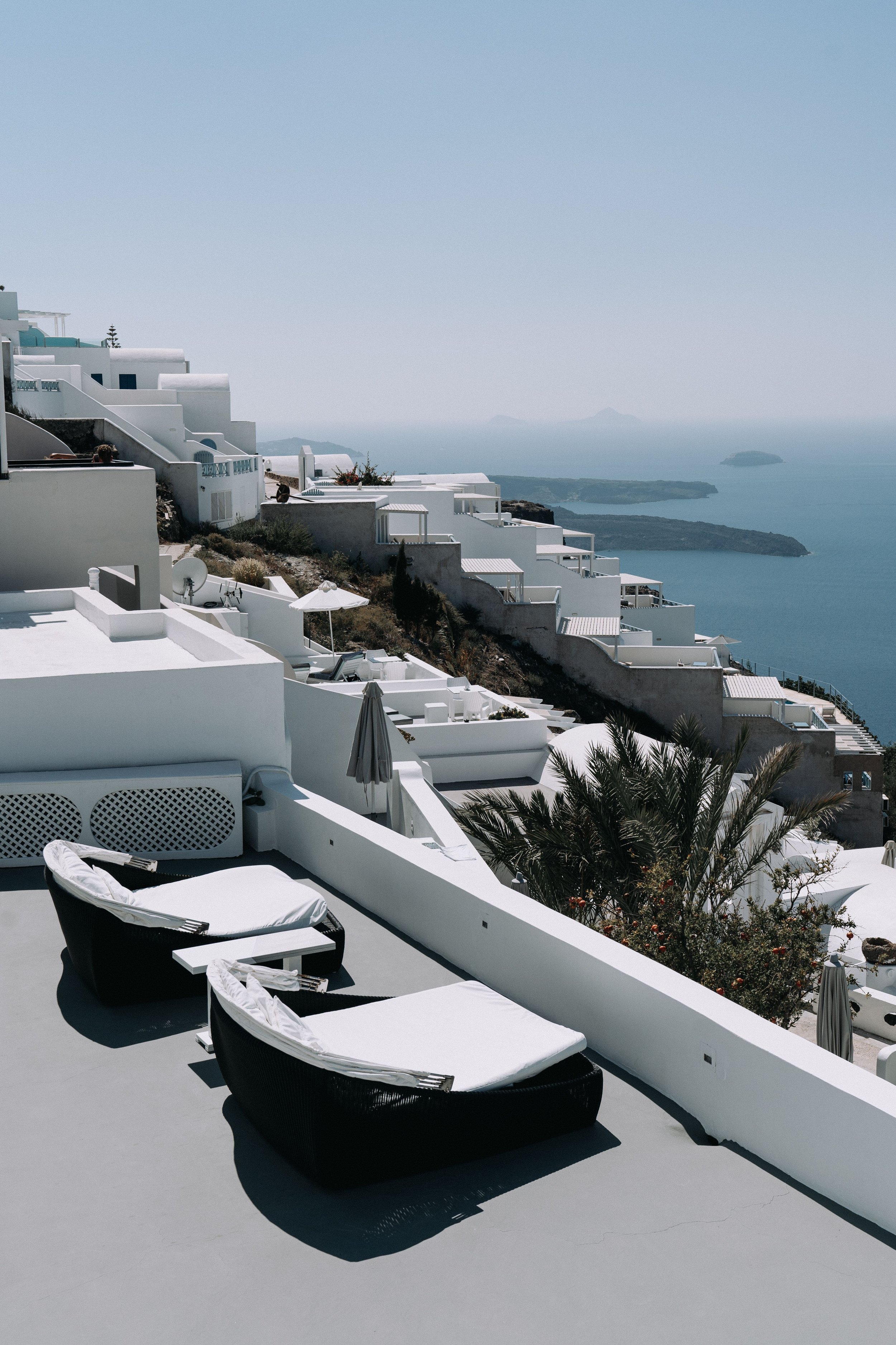 Beachside outdoor area with white loungers, umbrellas, and buildings on a hillside overlooking the sea and islands in the distance.