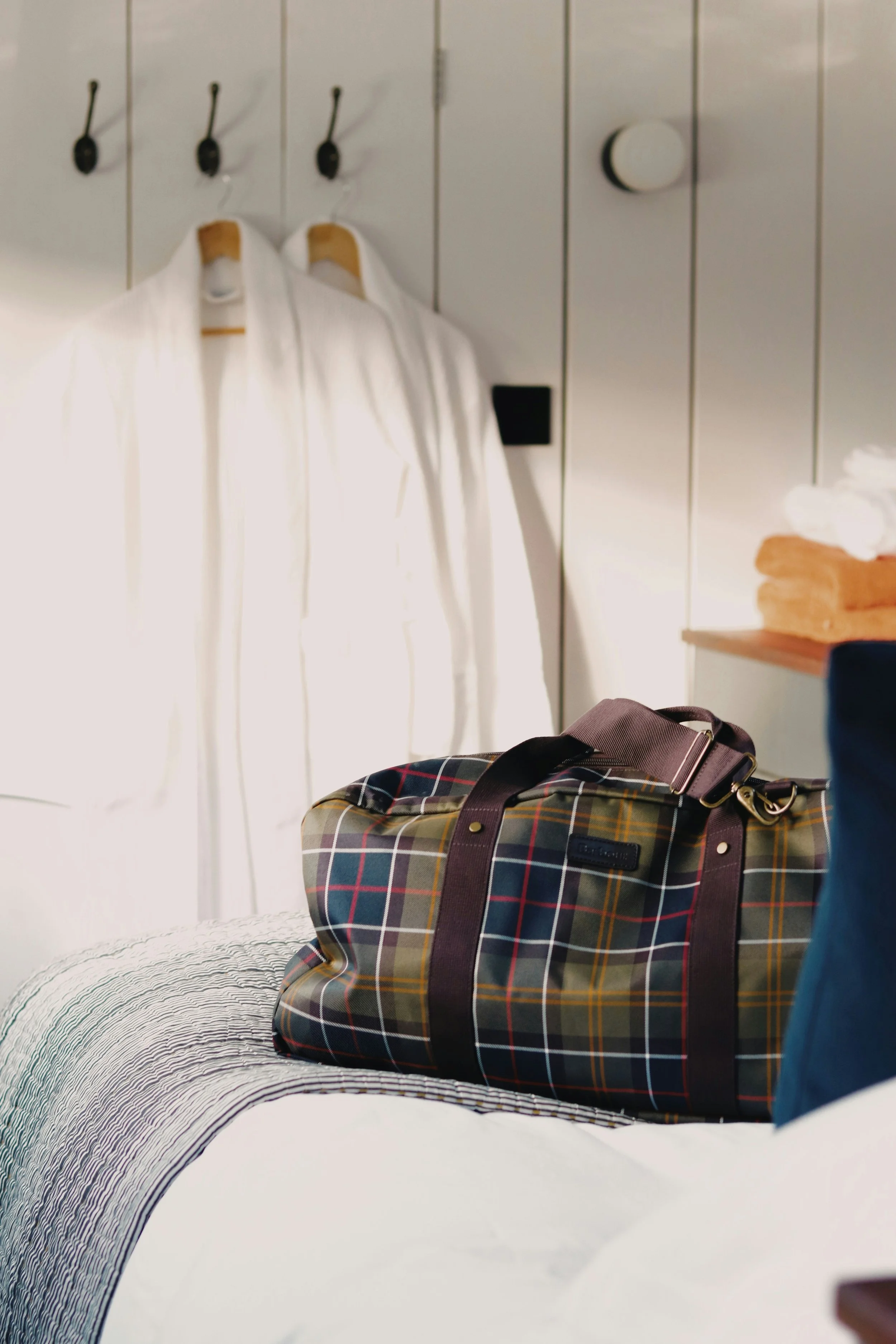 A hotel room with white wooden panel walls, containing white bathrobes hanging on hooks, a luggage bag with a plaid pattern on the bed, and some folded white towels on a wooden shelf.