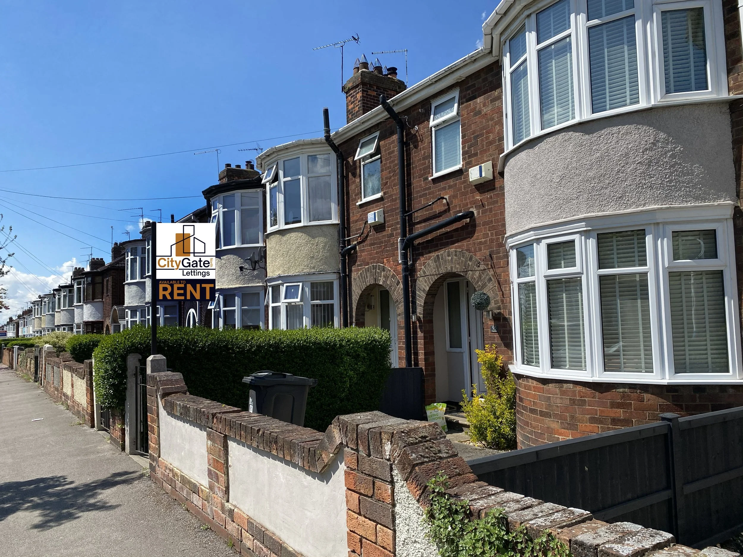 Street with a red "For Sale" sign in front of a brick residential house, featuring classic Victorian architecture, and adjacent greenery on a cloudy day.