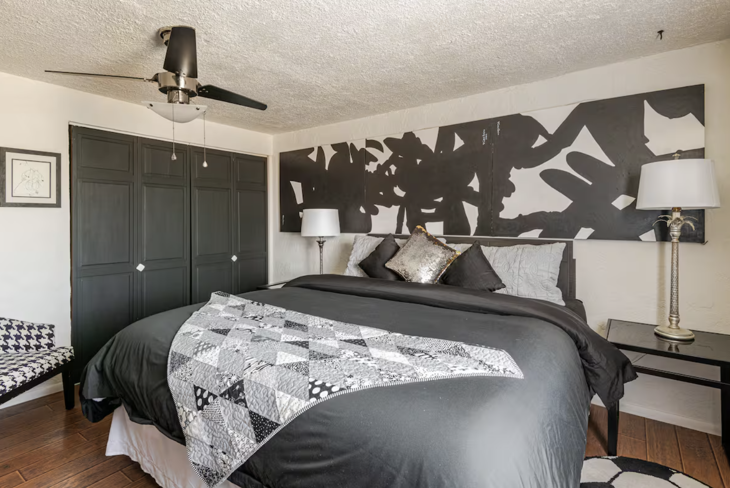 A bedroom with a black and white color scheme, featuring a large bed with multiple pillows and a geometric quilt, black closet doors, abstract black and white wall art, a black nightstand with a silver lamp, a bench with houndstooth pattern, ceiling fan, and hardwood flooring.
