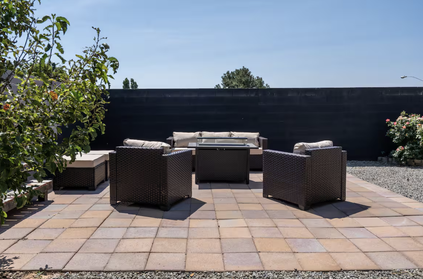 Outdoor patio with black wicker furniture, including a sofa, chairs, and a small table, on paving stones, with shrubbery and flowers against a dark fence and a clear blue sky.