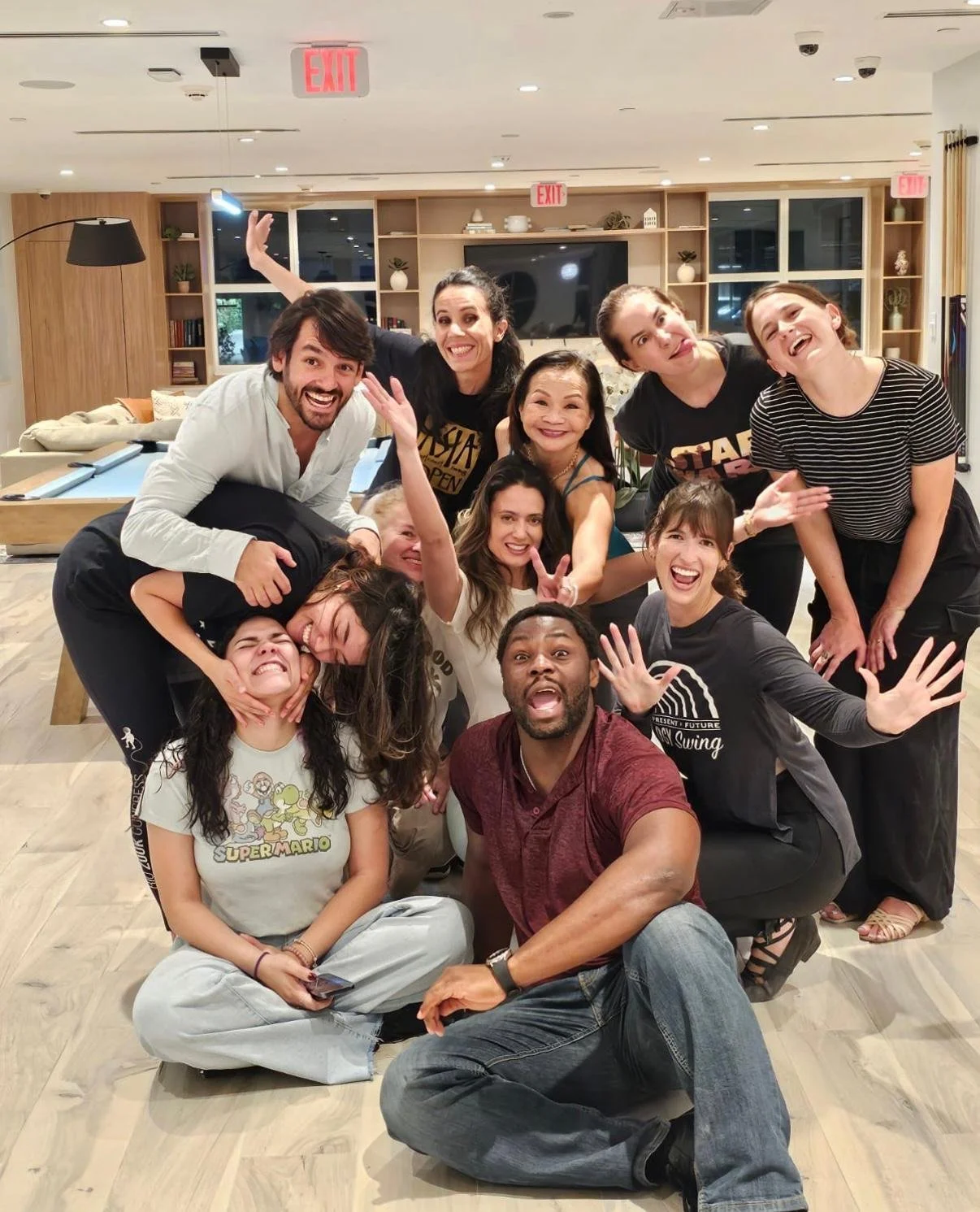 A group of ten people in a living room, smiling and making playful gestures for a photo. The room has wooden shelves, a television, and a pool table in the background.