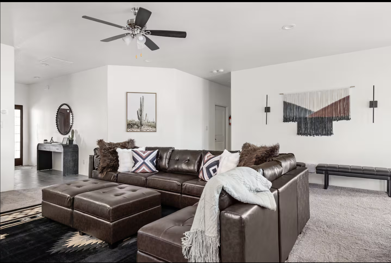 Living room with a brown leather sectional sofa, decorative pillows, and a matching ottoman. Wall art includes a cactus picture and a woven wall hanging. A black bench is positioned against the wall, and a ceiling fan with lights is on the ceiling. There's a doorway with glass panels and a small console table with decor near the entrance.