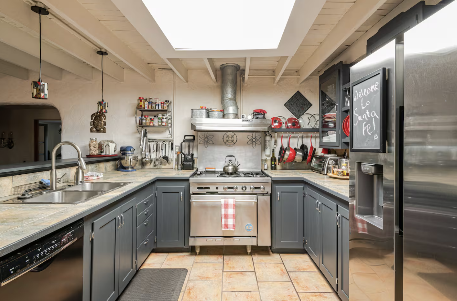Kitchen with gray cabinets, stainless steel stove, black refrigerator with chalkboard writing, tile countertops, and various kitchen utensils and appliances.