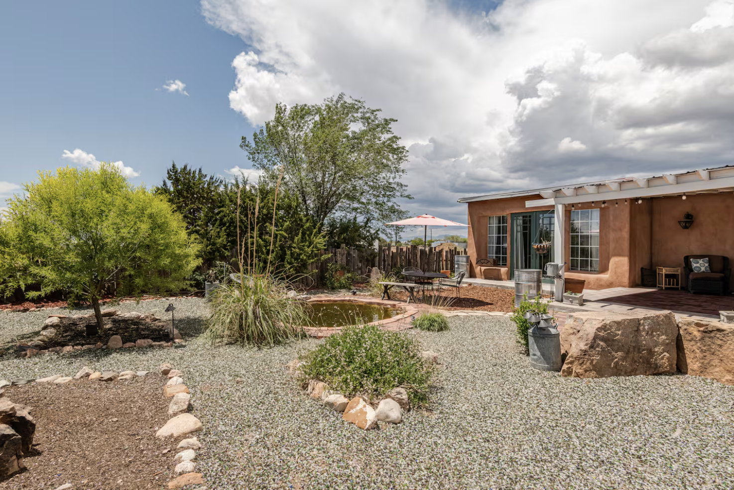 A backyard with a small pond, green bushes, and trees, surrounded by rocks and gravel. There is outdoor furniture including a table, chairs, and an umbrella near the house, which has a patio area with large windows and a sliding door. The sky is partly cloudy.