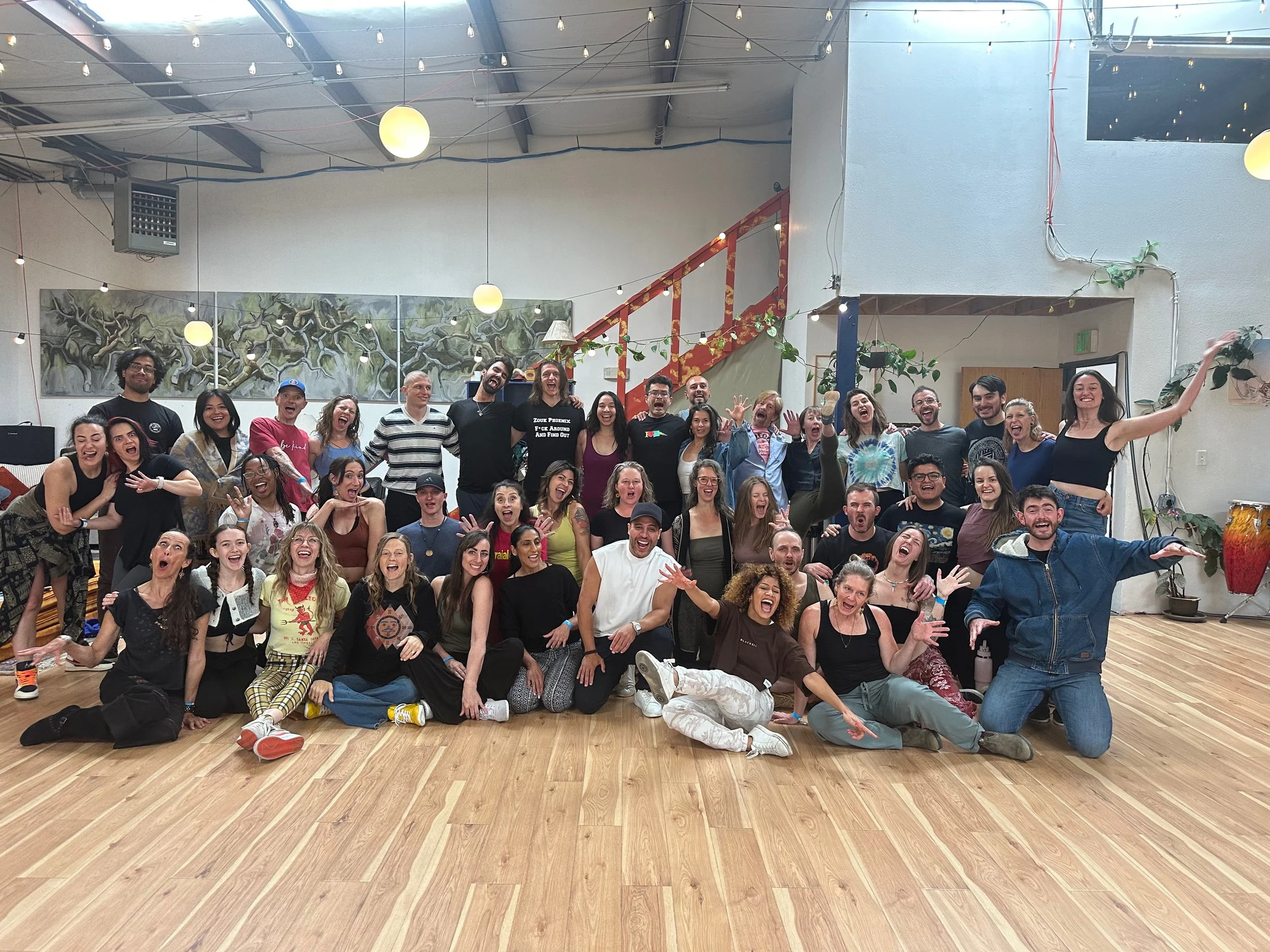 A large group of people is gathered indoors, smiling, laughing, and posing for a group photo. They are diverse in age and appearance. The room has a high ceiling with string lights, some artwork on the walls, and a wooden floor. The atmosphere appears joyful and lively.