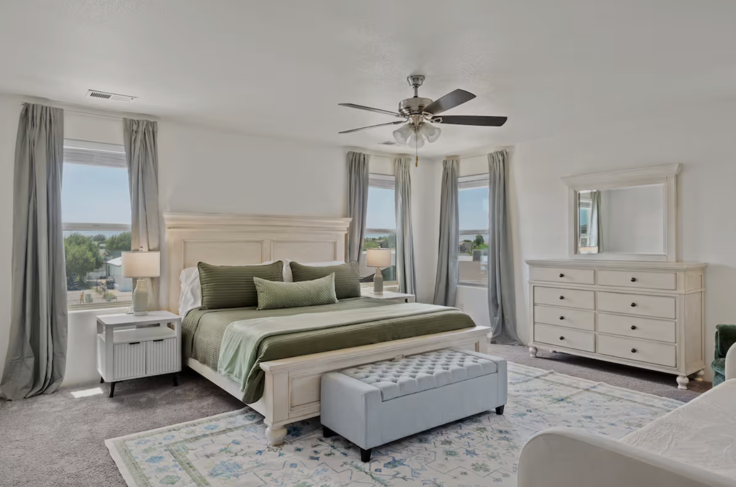 A bright bedroom with a large bed, green and white bedding, and matching pillows. There is a white dresser with a mirror above it, a bedside table with a lamp, and windows with curtains that let in natural light. An upholstered bench is at the foot of the bed, and a ceiling fan is overhead.