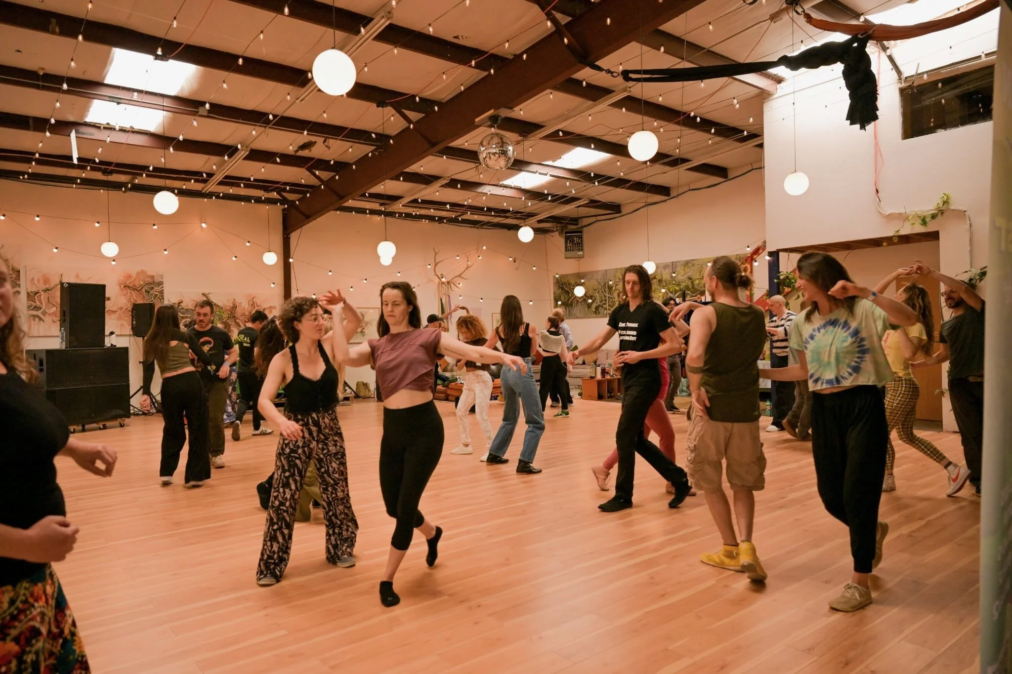 People dancing in a spacious indoor dance floor decorated with string lights, globe lights, and a disco ball, with wooden flooring and artistic displays on the walls.