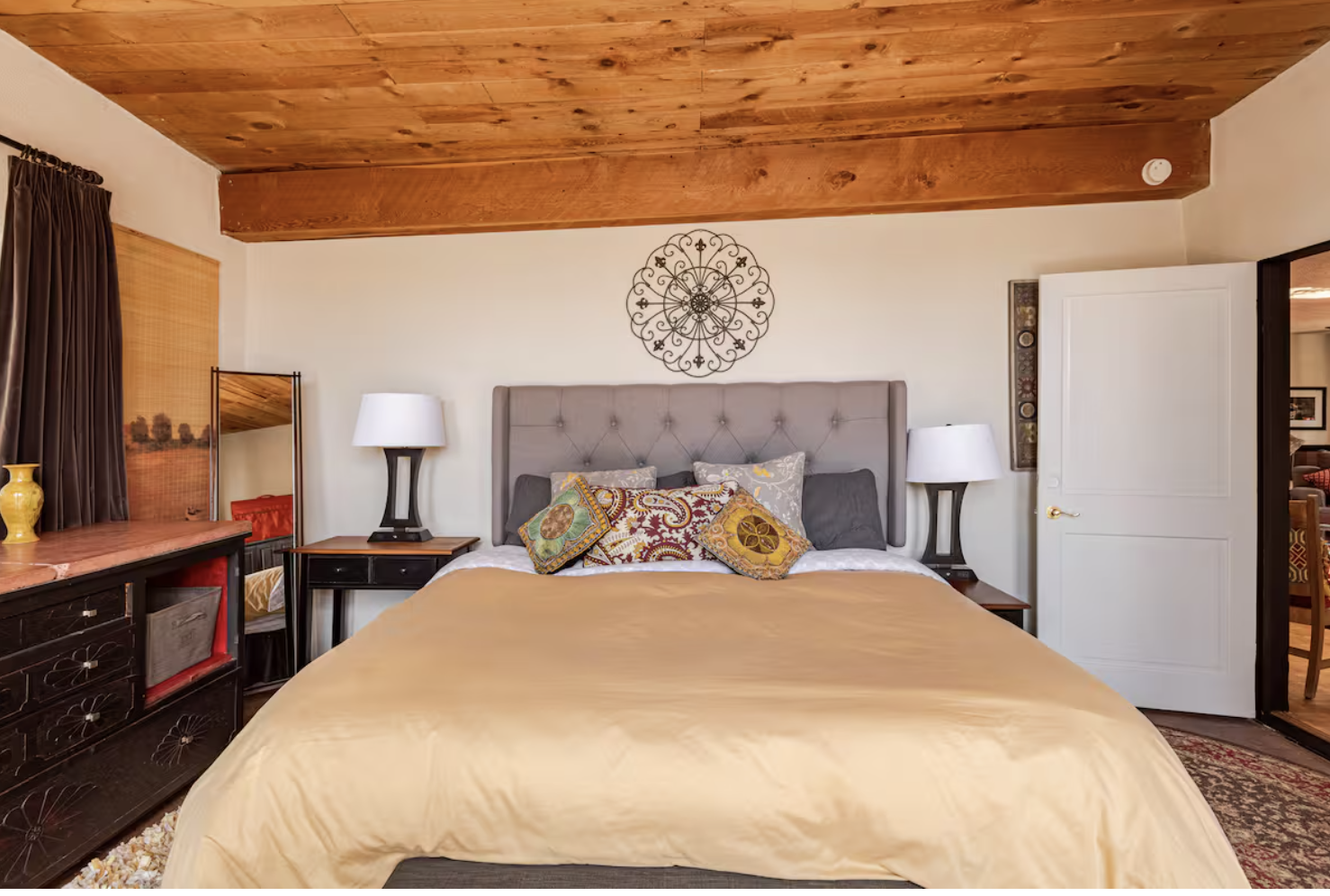 A bedroom with a wooden ceiling, beige walls, and a large bed with a tufted headboard adorned with decorative pillows. Two nightstands with lamps flank the bed, and a mirror, a window with dark curtains, and a dresser are visible in the room.