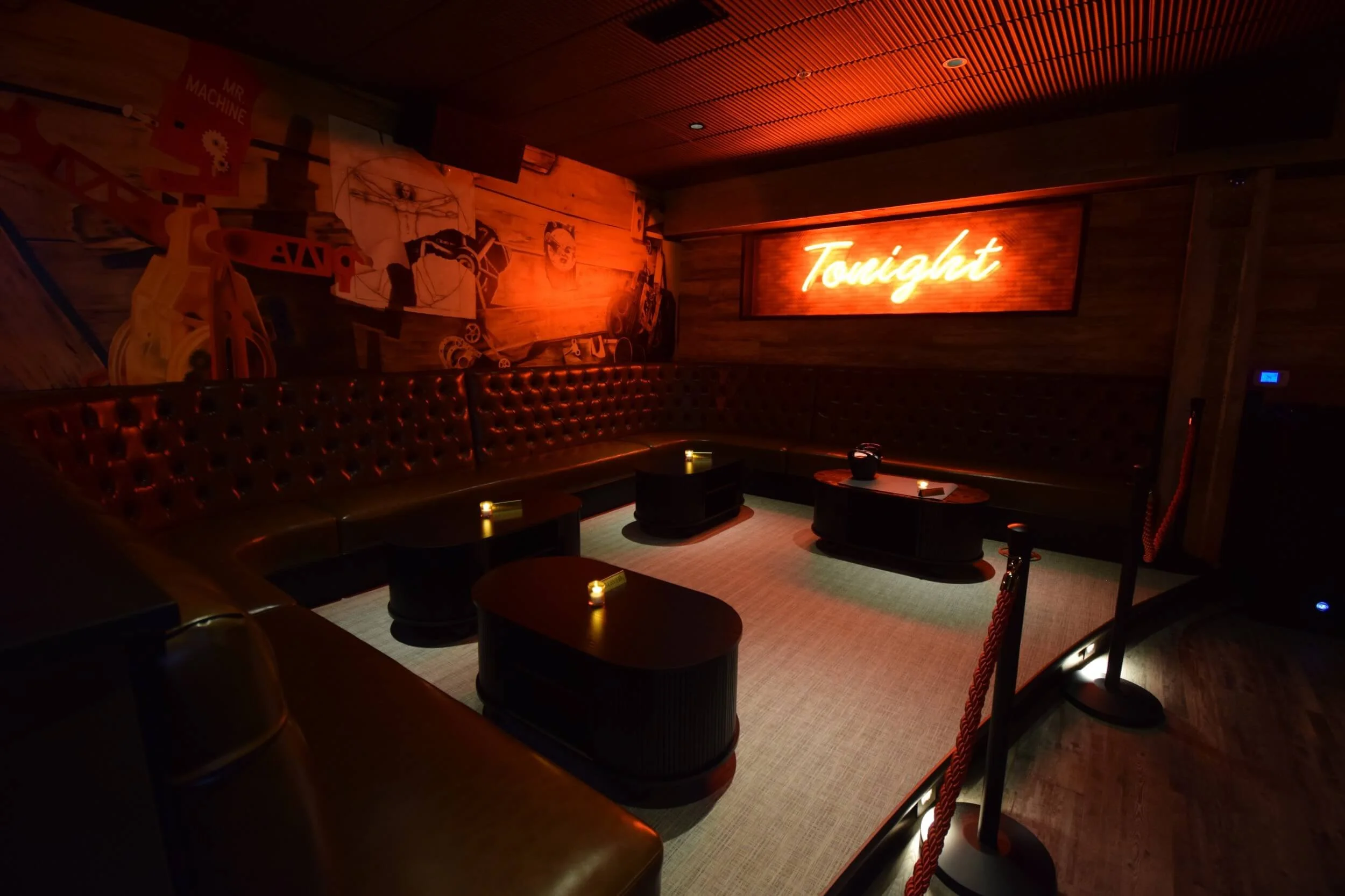 Dark, cozy lounge area with leather seating, small tables, dim lighting, and a neon sign that says 'Tonight' on a wood-paneled wall, with artwork on the walls.