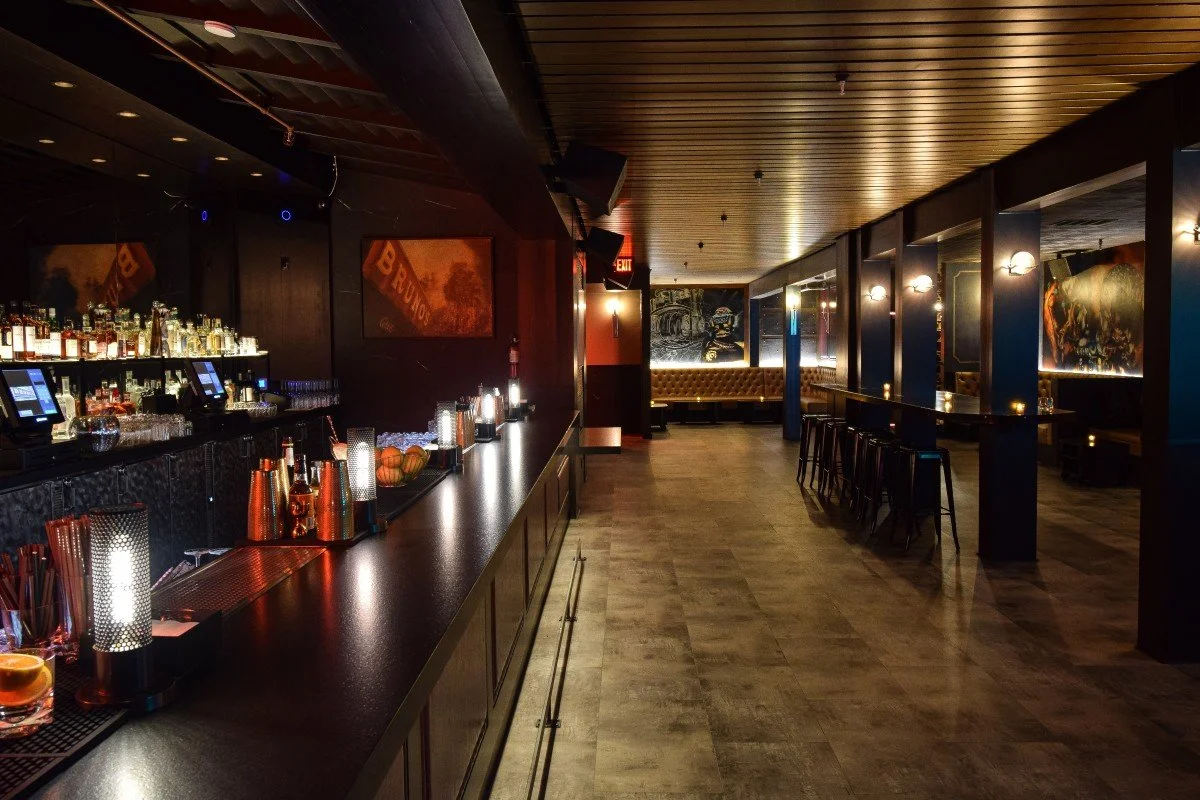 Elegant bar with a long black countertop, bottles of liquor and glasses, and bar tools. There are bar stools along the counter, artwork on the walls, and warm lighting.