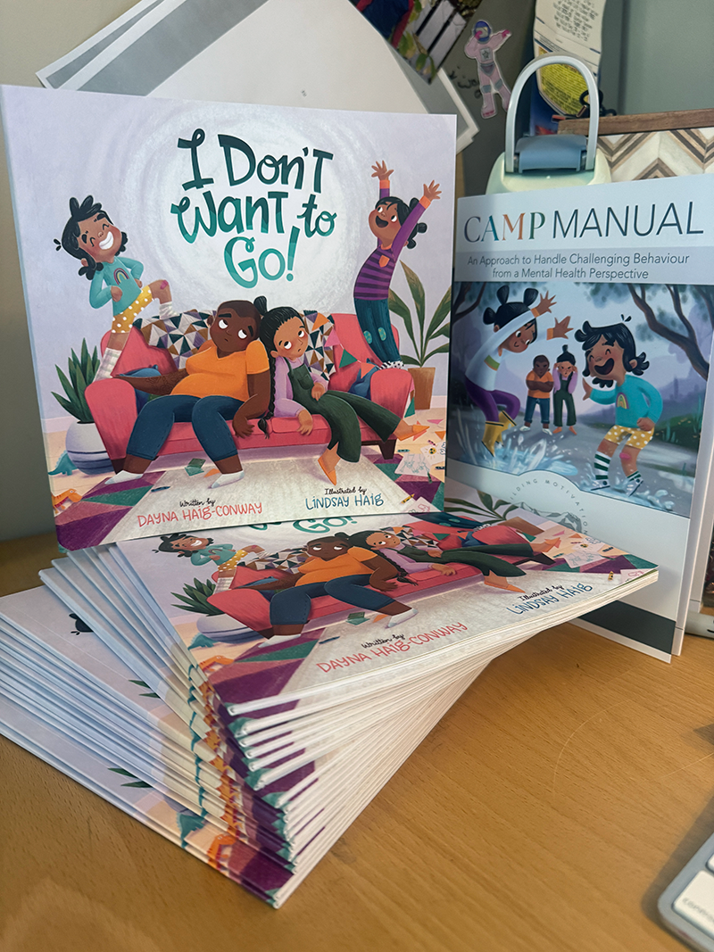 Multiple copies of a children's book titled 'I Don't Want to Go!' stacked on a wooden surface, with additional books and materials related to camping and challenging behavior in the background.