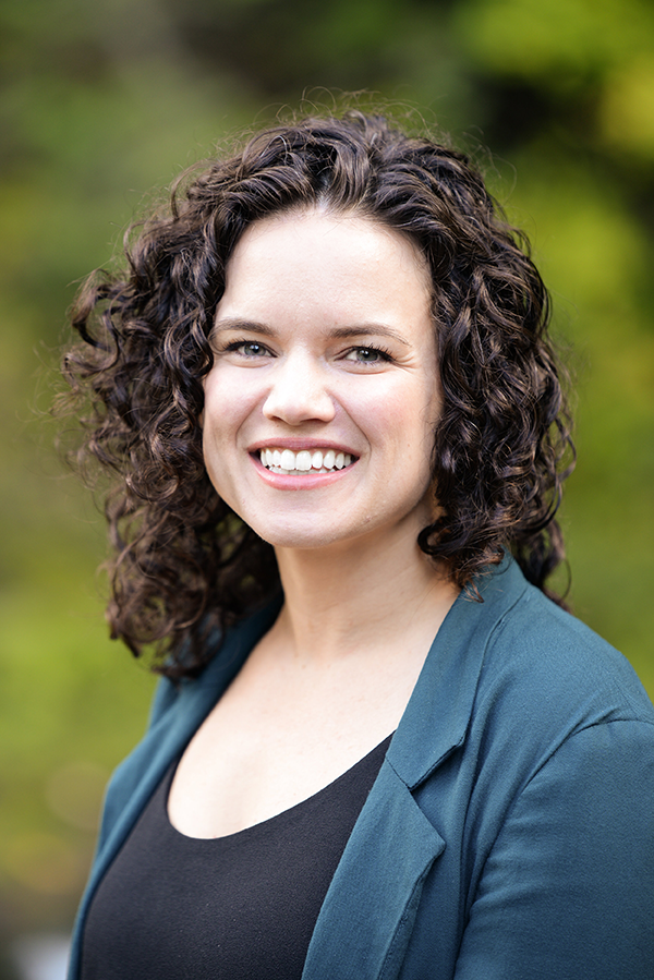 Dr. Kallie Doucette, ND (MSCP) hair smiling outdoors in front of green trees, wearing a black top and teal blazer.