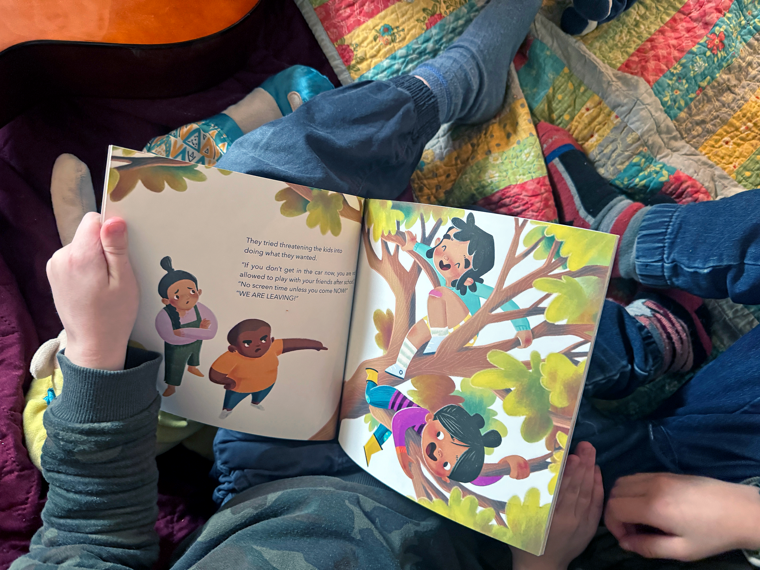 A child sitting on a quilted blanket, holding an open children's book, 'I Don't Want to Go," with colorful illustrations of children climbing a tree, while an adult reads along.