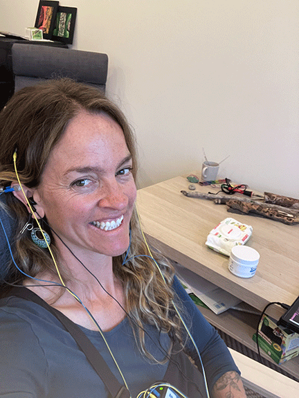Dayna, a woman with long hair taking a selfie in a room, smiling, with various objects on a table in the background, including tools, a small container, and a box.