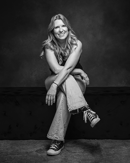 Dr. Tyla Mackay on a black bench against a dark background, smiling and wearing casual clothing including sneakers.