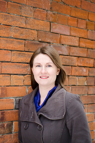 Anne Keery with a blue shirt standing in front of a brick wall.