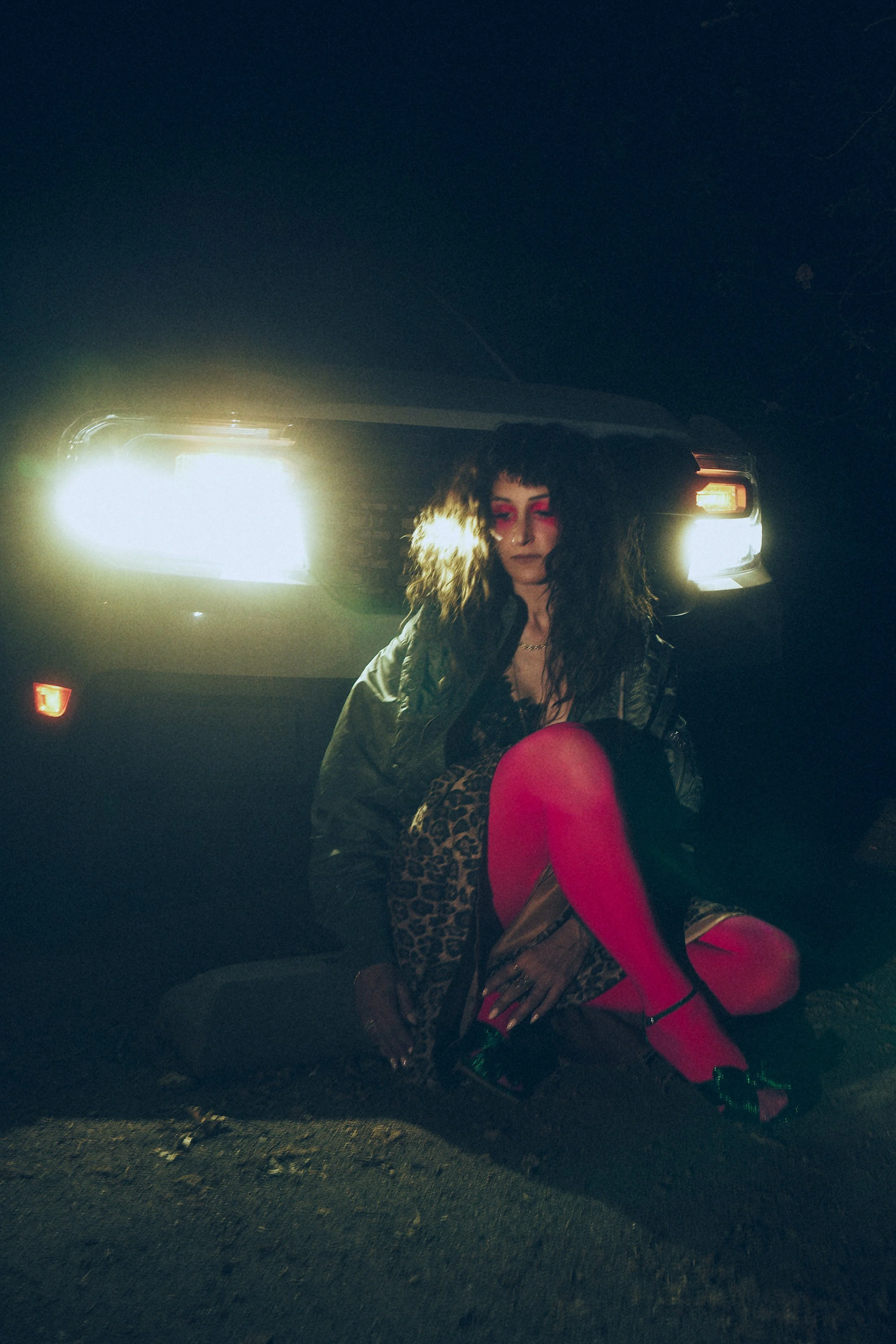 Tracy Marcellino AKA Witch Cabin with long, curly hair, wearing bright pink stockings, high heels, a black leather jacket, and leopard print clothing, sitting on the ground in front of a car with its headlights on at night.