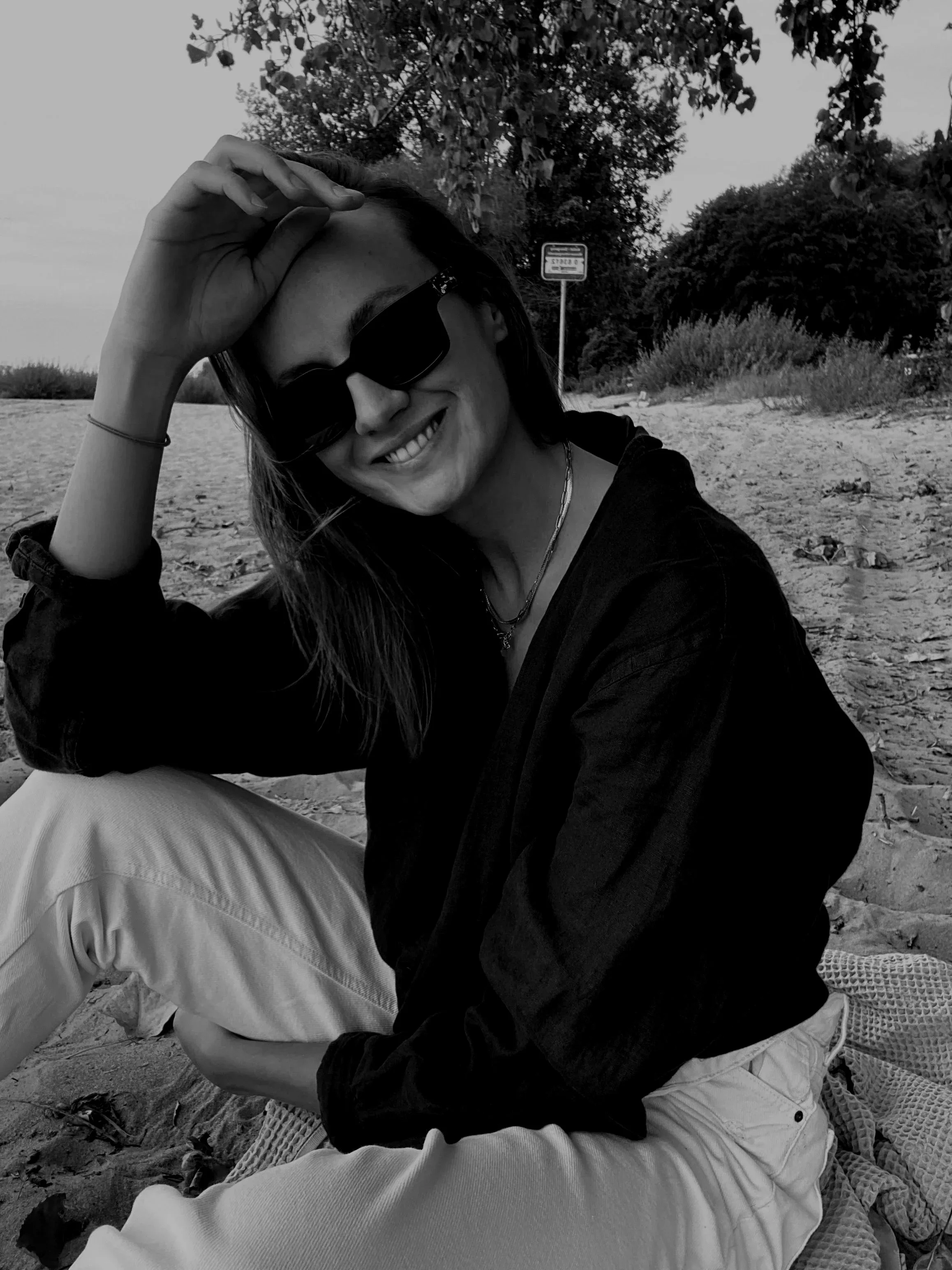 A woman with long hair, wearing sunglasses, smiling, sitting on a beach with trees and a sign in the background.