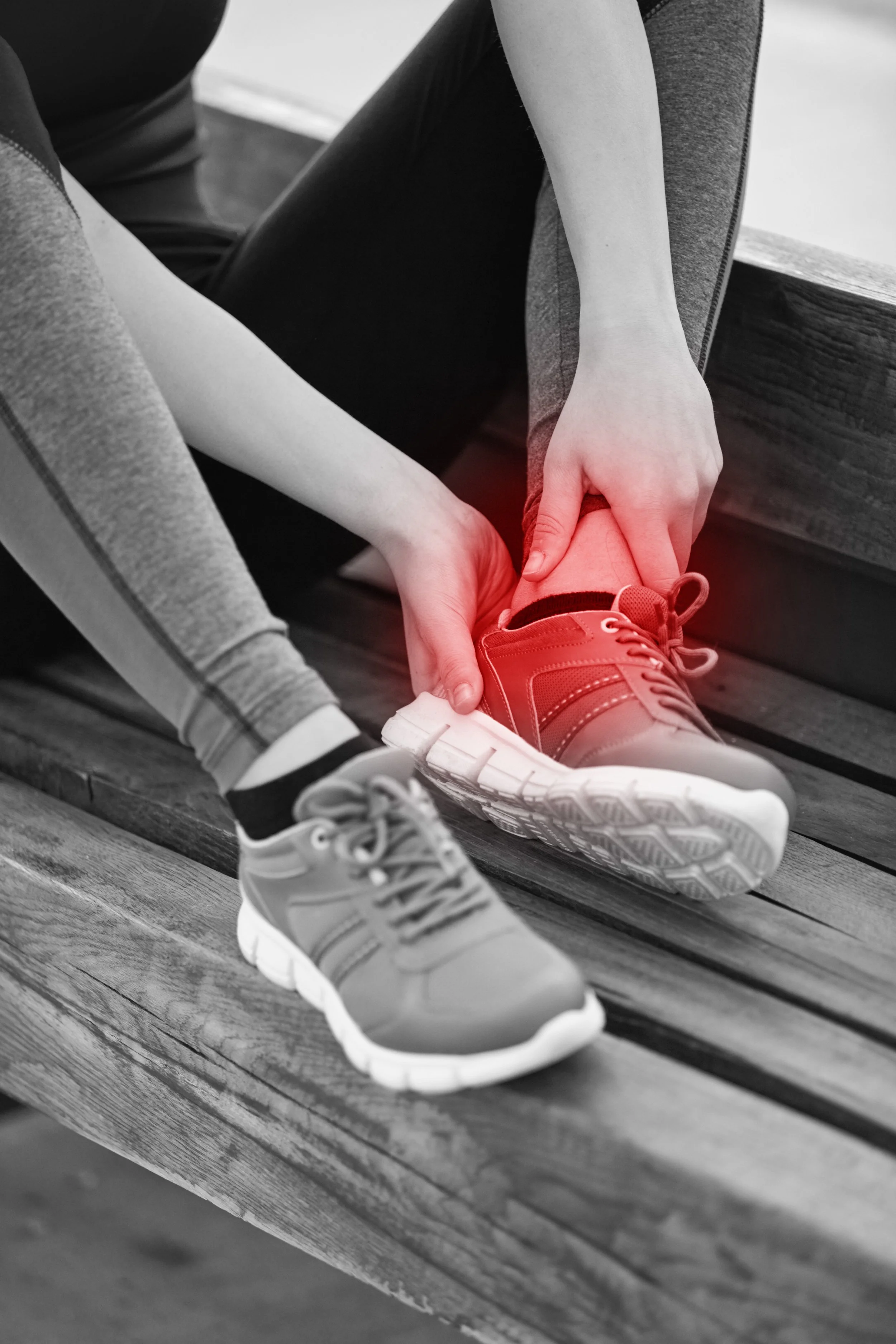 A person helping another person put on a red sneaker on a wooden bench, with the rest of the image in black and white.