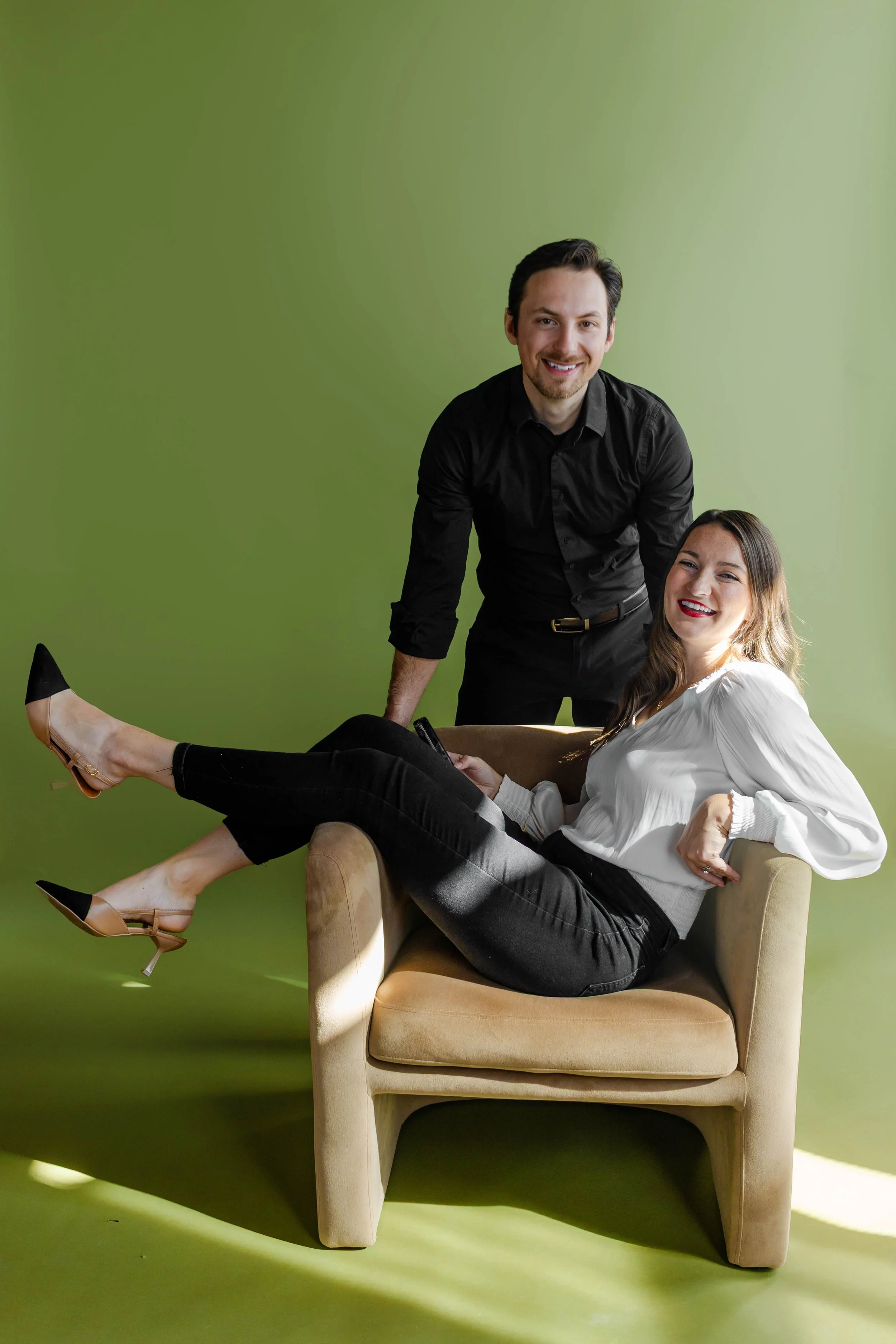A man and a woman smiling, with the woman sitting on an armchair with her legs extended onto the floor, against a green background.