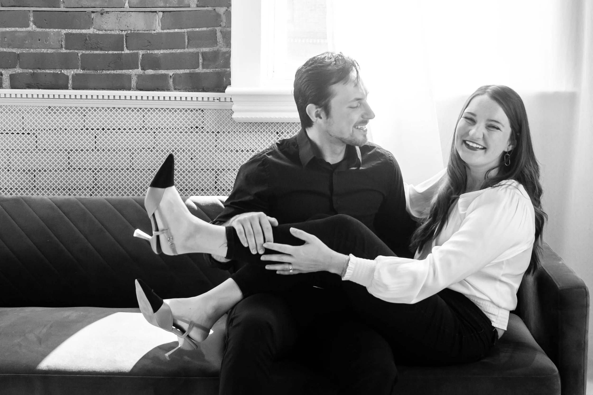A man and woman sitting on a sofa, smiling and laughing, with the woman holding the man's leg. The image is in black and white.